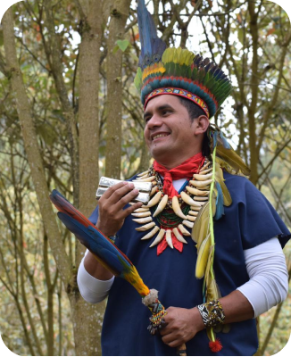 An indigenous man in traditional attire, wearing a vibrant feathered headdress and a large bone necklace, smiling while holding a small flute in a forest setting