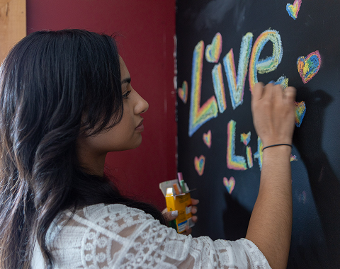 A person with long dark hair writes "Live Life" in colorful chalk on a blackboard adorned with rainbow hearts.